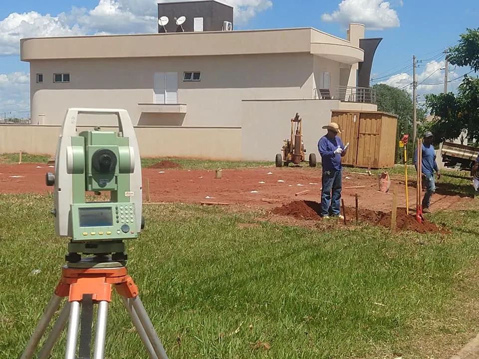 Estação total em tripé no gramado, com a equipe trabalhando diante de um sobrado em obra