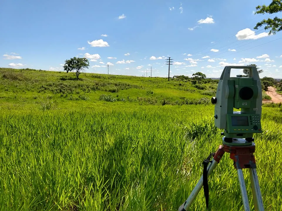 Estação total sobre tripé num campo de capim verde, com céu azul e uma árvore ao fundo